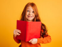 Bücher begeistern – Vorlesewettbewerb Jahrgang 6 Image of cheerful little redhead girl with freckles standing isolated over yellow background reading book. Looking camera.