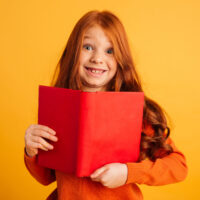 Bücher begeistern – Vorlesewettbewerb Jahrgang 6 Image of cheerful little redhead girl with freckles standing isolated over yellow background reading book. Looking camera.